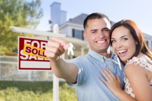 Couple In Front of New Home with New House Keys and Sold Real Estate Sign Outside.